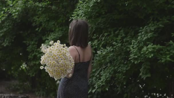 Νεαρό κορίτσι πηγαίνει στο βάθος με ένα μπουκέτο λευκό χαμομήλι στο δρόμο ενάντια στο πράσινο του πάρκου — Αρχείο Βίντεο