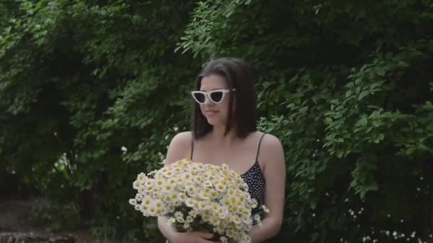 Όμορφη νεαρή γυναίκα με γυαλιά και ένα μπουκέτο λευκά λουλούδια από μαργαρίτα χαμομηλιού στο δρόμο στο πάρκο κοντά στο πράσινο θάμνο — Αρχείο Βίντεο