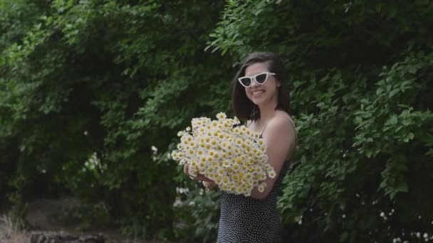 Joven hermosa mujer bonita con gafas y un ramo de flores de margarita de manzanilla blanca en la calle en el parque cerca del arbusto verde — Vídeos de Stock