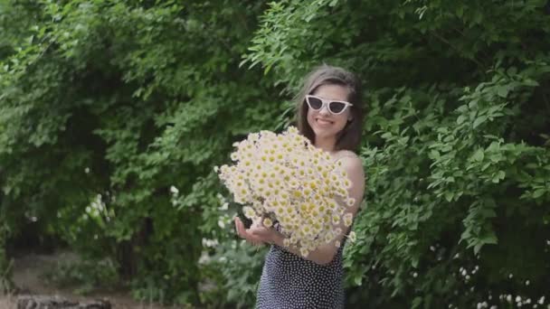 Wanita cantik muda dengan kacamata dan karangan bunga bunga bunga bunga daisy chamomile putih di jalan di taman dekat semak hijau — Stok Video