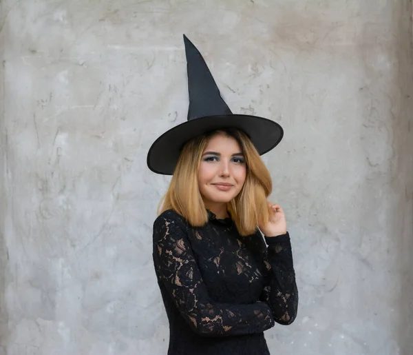 Mujer joven gótica feliz en traje de Halloween bruja con sombrero de pie y sonriendo sobre fondo gris —  Fotos de Stock