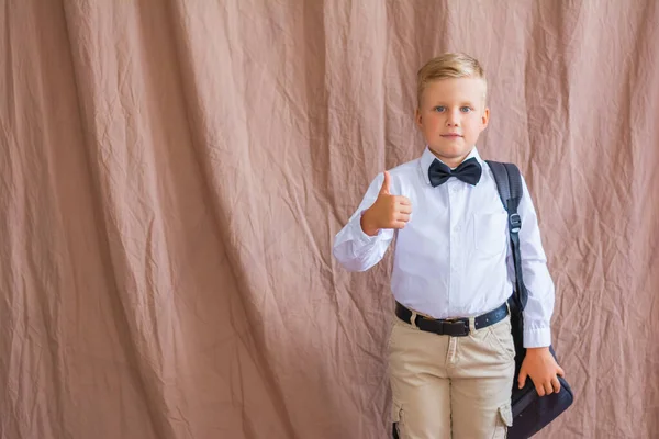 Retrato de un apuesto colegial sonriente con una mochila mostrando como en una camisa blanca y pantalones beige con una pajarita —  Fotos de Stock