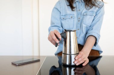 Woman holding coffee pot in the kitchen of her home, on the counter top is her mobile phone