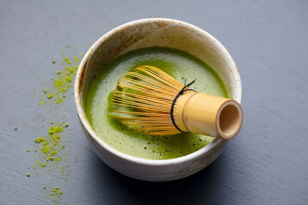 Matcha green tea cooking process in a bowl with bamboo whisk. Grey background. Close up.