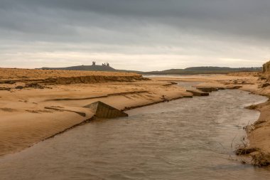 Northumberland, İngiltere 'deki Dunstanburgh Şatosu, nehir önplanda gezinirken ve gökyüzü bulutluyken çekildi.