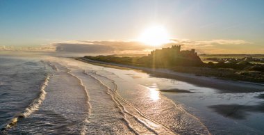 Güneş doğarken dondurucu soğuk bir sabahta Northumberland, İngiltere 'de karla kaplı kumsalın yanında insansız hava aracı tarafından çekilen ikonik Bamburgh Şatosu..
