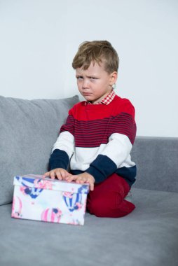 Upset boy sits with a gift on the bed.  clipart