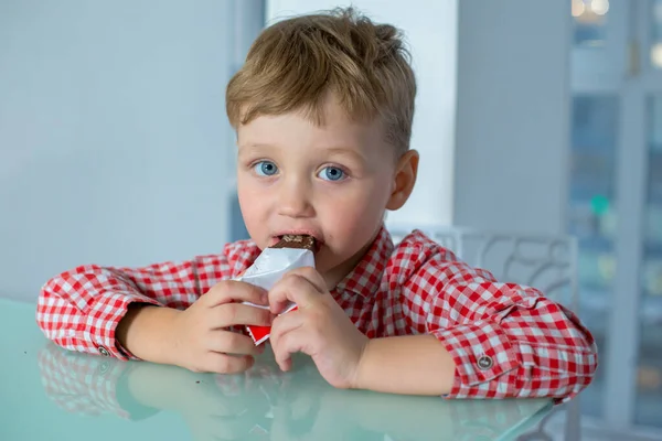 El chico se sienta a la mesa y se come una barra de chocolate. —  Fotos de Stock