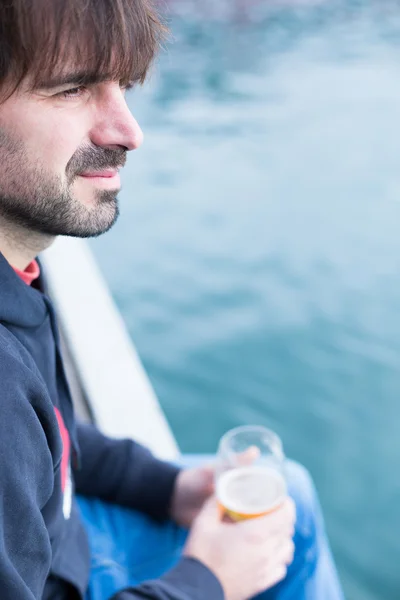 Brodaty mężczyzna siedzi w pobliżu wody i trzymając szklankę piwa — Zdjęcie stockowe