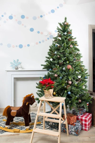 クリスマスツリーの背景に工芸品の灰色の紙に包まれた鍋にPointsettia 白い壁の背景に暖炉 壁にガーランドとロッキングホース — ストック写真