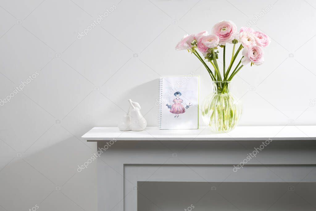 Bouquet of pink Persian buttercups in a glass figured vase on a white fireplace console. A faience figurine of two pears and a child's drawing as an interior decoration. Copy space