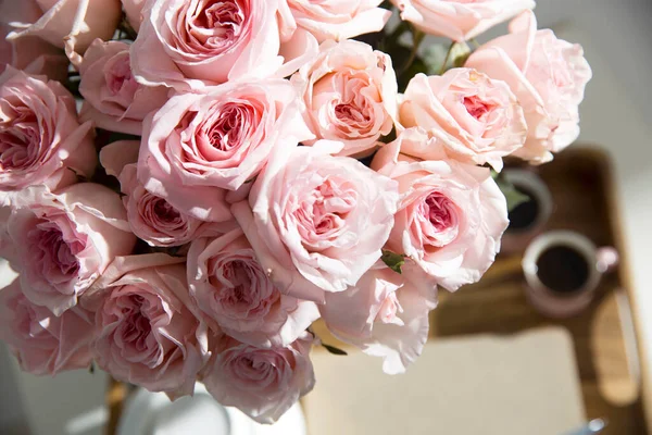 Rose White Pink O\'hara. Bouquet of pink roses in a glass vase with a white teapot and two cappuccino cups on a tray on a coffee table among books.