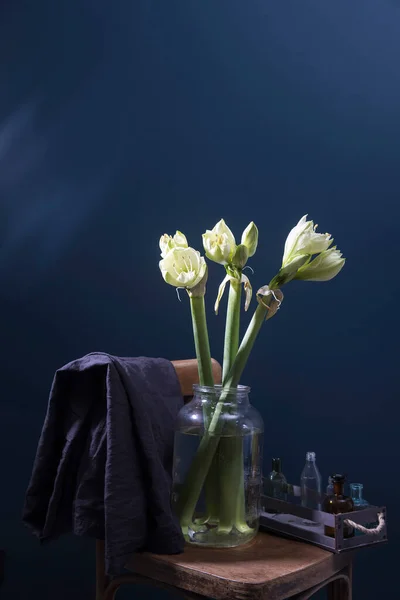 一束未吹灭的绿色和白色的芳纶花 放在玻璃瓶中的维也纳椅子上 靠着一面深蓝色的墙 小药瓶的木制托盘 椅子的后面挂着一块灰色的布 — 图库照片