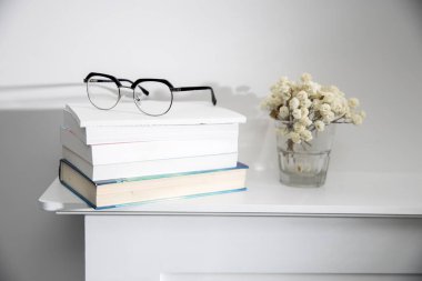 Bir yığın kitap, üzerinde gözlükler, beyaz bir masanın üstünde. Yakınlardaki cam bir vazoda kurutulmuş çikila çiçekleri. Boşluğu kopyala