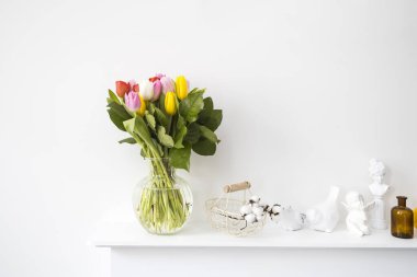 Bir buket çok renkli lale, içinde pamuk çiçekleri olan bir hasır metal sepet. Bir kuş heykeli, dişi bir göğüs sahte bir şöminenin panelinde duran iki peri kuşu. Kahverengi eczane şişesi.