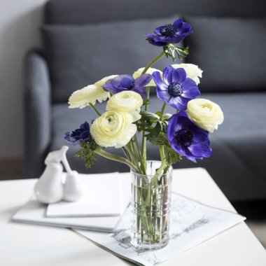 Beyaz ranunculus buketi ve mavi şakayık, ekoseli kanepenin yanındaki beyaz sehpanın üstünde. Kitaplar ve günlük. Gölge. Kare kare