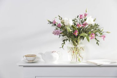 Beyaz lalelerden oluşan minimalist bir buket pembe Eustoma, sümbül, okaliptüs flütlü cam bir vazoda yapay bir şömine panelinde. Çay fincanı ve çay tabağı, bir kuş ve bir meleğin heykeli. Boş boşluk.
