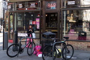 Londra, İngiltere - 20 Eylül 2020, Brick Lane 'deki barın yakınına bisikletler park edilmiş. Kız telefonuna bakıyor. Çöp kutusu dolu. Çöp torbaları çöp kutusunun yanında.