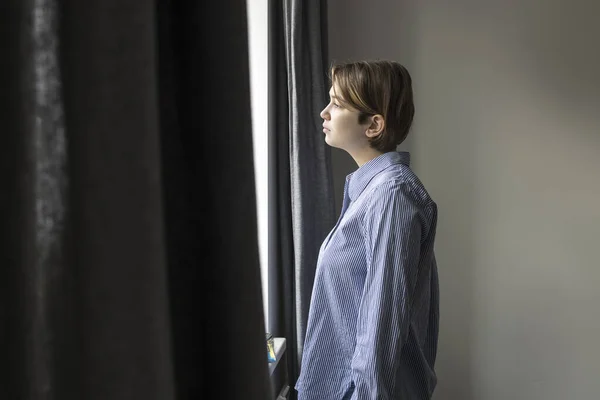 一个有着19岁短发 穿着蓝色条纹白衬衫的沉思女孩从窗户向外张望 灰色窗帘 — 图库照片