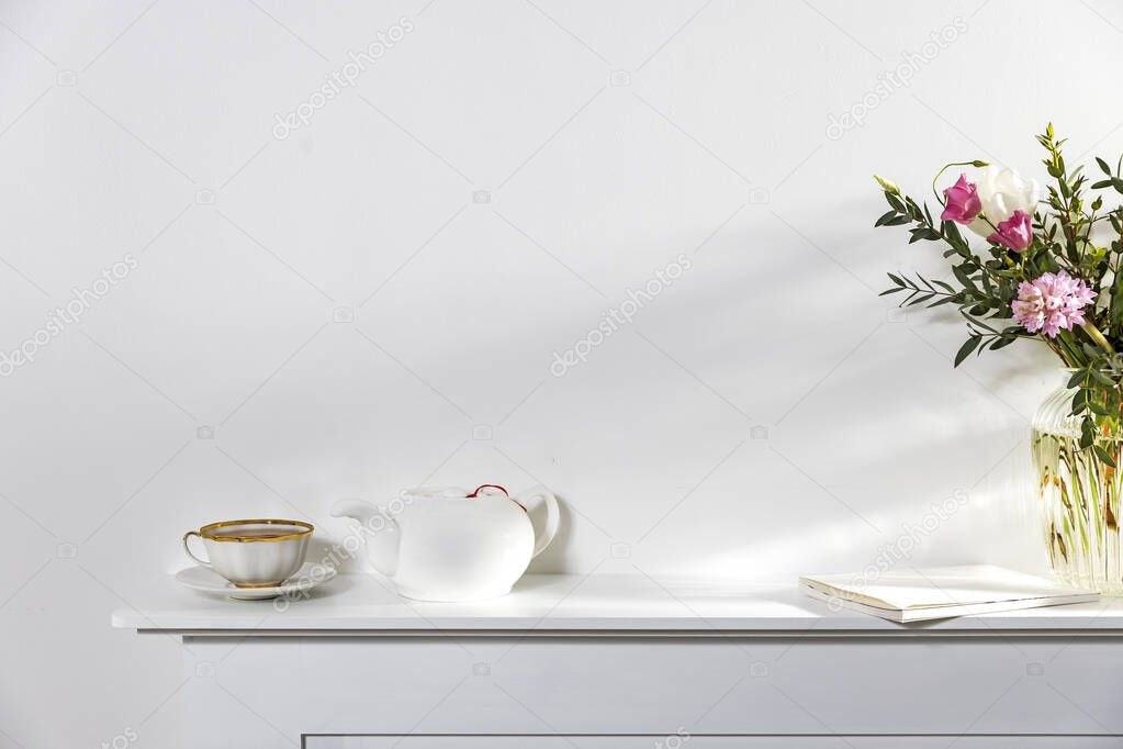 White on white. A cup of tea, a kettle, and a notebook stand on a white panel of an artificial fireplace. Empty space.