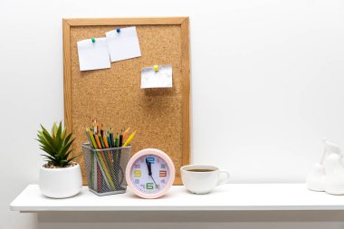 Üzerinde çivilenmiş kağıtlar olan bir not tahtası, seramik kapta yapay sulu, pembe çalar saat, beyaz masa üstünde kurşun kalemler olan bir kalem tutacağı. Boşluğu kopyala