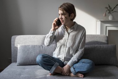 Mavi çizgili beyaz gömlekli, kısa saçlı, düşünceli bir kız telefonda konuşuyor, gri kanepede yastıklarla gülüyor. Yatay çerçeve