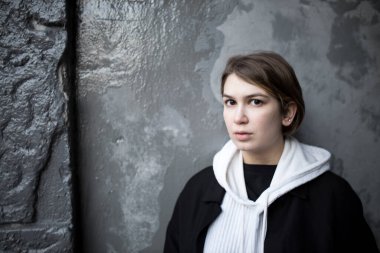 Beyaz kapüşonlu, siyah yağmurluk ve kot pantolonlu, kısa saçlı, ciddi bir kız sokakta yürüyor. Saçları boyanmış..