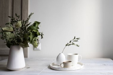 Minimalist İskandinav tarzı. Beyaz flütlü bir vazoda elma ağacının yarı açık dalları. Kurumuş okaliptüs dallı küçük bir vazo ve masanın üzerindeki tepside iki fincan kahve..