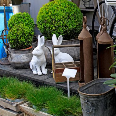 Boxwood çalıları ve bahçe çimenleri arasında iki seramik tavşan. Tekerlekli tramvay ve çeşitli bahçe aletleri - fenerler, ahşap sandıklar