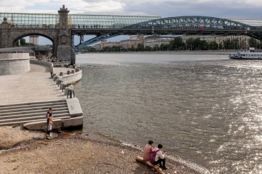Moskova, Rusya - 15 Mayıs 2021: Puşkin setinin manzarası. İnsanlar nehrin kıyısında kütüğe oturur. Gemi köprünün altından geçiyor. Anne ve çocuk merdivenlerde durun. Gorky Kültür ve Eğlence Merkezi Parkı