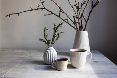 Minimalist İskandinav tarzı. Beyaz flütlü bir vazoda elma ağacının yarı açık dalları. Kurumuş okaliptüs dallı küçük bir vazo ve masanın üzerindeki tepside iki fincan kahve..
