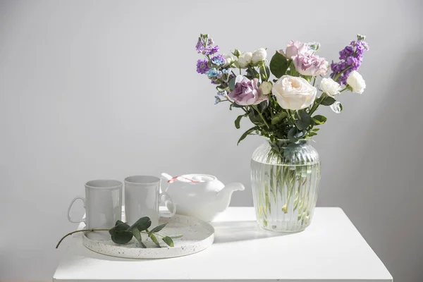 ハケリア ヴェルチナ 紫と白のバラ 小さなお茶のバラ マッティオラ インカーナとガラスの花瓶に青い虹彩の花束 内部の白いテーブルの上にユーカリの枝とトレイ上のティーポットと2つの背の高いティーカップ — ストック写真
