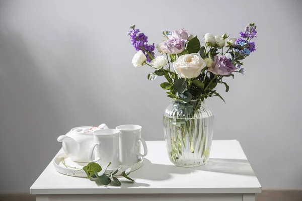 ハケリア ヴェルチナ 紫と白のバラ 小さなお茶のバラ 抹茶のインカーナとガラスの花瓶の青の虹彩の花束は 白いコーヒーテーブルの上にあります 灰色の壁 — ストック写真