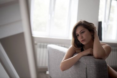 20 yaşında mayo giymiş esmer bir kız arkası pencereye dönük bir şekilde kanepede oturuyor.