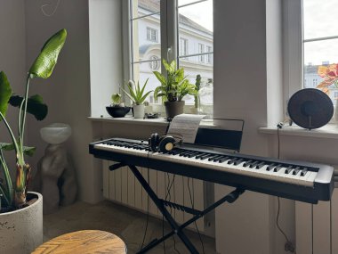 Varşova, Polonya, 28 Haziran 2025. Dijital piyano, kulaklıklar ve notalar ev bitkileriyle dolu parlak bir pencerenin yanında.