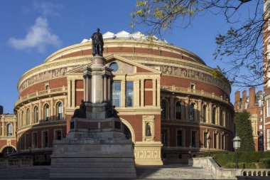 Londra, İngiltere, 2 Mayıs 2025. Kraliyet Albert Salonu 'nun heybetli, yuvarlak kırmızı tuğla mimarisi güneşli bir günde ön planda öne çıkan bronz bir heykel ve anıt tarafından domine edilir..