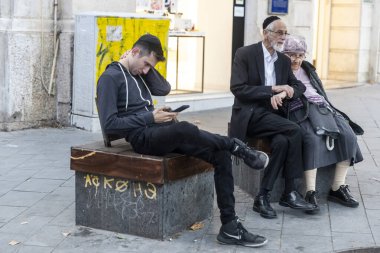 Kudüs, İsrail, 23 Ekim 2025, telefonuna takılmış genç bir Yahudi ile sokak mobilyalarının yanında oturan yaşlı bir dindar çift arasındaki zıtlık.