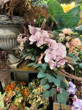 Pastel pembe güve orkideleri, tozlu mor ve kahverengi kuru elementler ve okaliptüs benzeri yeşillikler içeren özenle hazırlanmış, yapay bir çiçek aranjmanı yıpranmış bir kül vazosunun etrafında toplanmış..