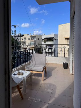 Yoğun inşa edilmiş bir şehir caddesine bakan bir apartman balkonunun manzarası. Bir sandalye, küçük bir masa ve beyaz bulutlu açık mavi bir gökyüzü..