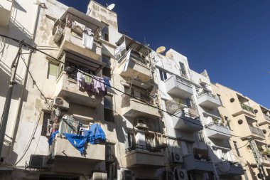 Birden fazla balkon, havalandırma ünitesi ve derin mavi bulutsuz bir gökyüzü altında korunmasız kablolar içeren, yakın bir şekilde paketlenmiş, yaşlanan apartmanların düşük açılı görüntüsü..