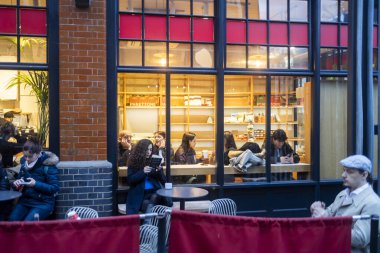 Londra, İngiltere, 23 Kasım 2025 'te, müşterilerin ahşap masalarda oturdukları, kitap okudukları veya telefonlarını kullandıkları, mola verdikleri sıcak ışıklı bir kafeye açılan büyük camdan bir manzara..