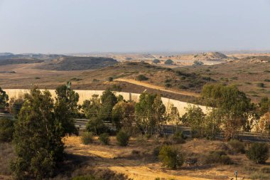 Kuru ve yuvarlanan manzaranın yüksek görünümü ön plandan geçen bir otoyol ve orta zemindeki tepeler boyunca kıvrılarak hassas sınır bölgesini işaret eden uzun ve yüksek beton bir güvenlik bariyeri gösteriyor. İsrail, Sderot