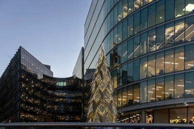 London, UK - December 1, 2024, A contemporary, geometric christmas tree made of lights standing prominently between two curved, modern glass office buildings in a city square at twilight.