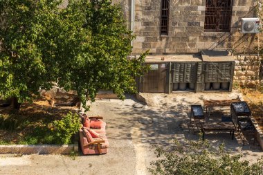 jerusalem, Israel, 2 November 2025, High-angle view of a simple, dusty backyard area next to a stone building, featuring a pink outdoor sofa, barbecue grills, and a large shade-casting tree.