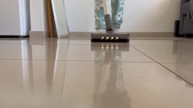 Açık yeşil pantolonlu ve beyaz çiçekli bir insan, açık fayanslı zemini yansımalarla süpürüyor. Mutfak temizliği. Kamera yere yerleştirilir ve vakum doğrudan kameraya doğru hareket eder..