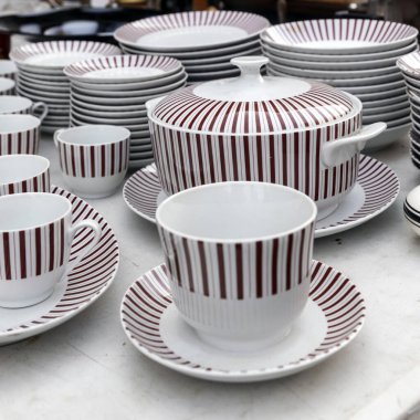 Tabaklar, çay fincanları ve bir çorba kabı da dahil olmak üzere dikey bordo şeritli beyaz porselenler, bir masanın üzerine yakın bir şekilde dizilmiş, desen ve bağ bozumunun miktarını vurguluyor..