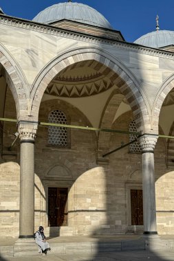 Türkiye, 5 Mayıs 2025 'te Süleyman Camii' nin güneşli avlusunda mermer kolonlar, çizgili kemerler ve etrafındaki kubbelerle bir kadın oturuyor. Ilık ışık, taş duvarlarda ve kapılarda büyük gölgeler yaratır..