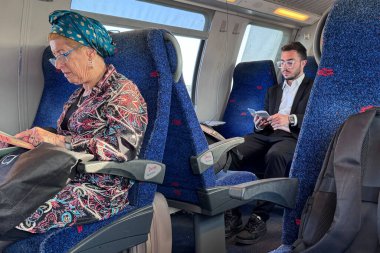 Trenle seyahat eden çeşitli yolcular, kitap okuyan yaşlı bir kadın ve telefonunu kontrol eden takım elbiseli genç bir adam da dahil.