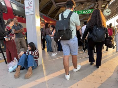 Tel Aviv, İsrail, 3 Kasım 2025, yolcularla dolu tren istasyonu sahnesi ve kırmızı tren vagonunun yanında oturan bir kız.