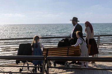 Hayfa, İsrail, 5 Ağustos 2025, güneşli bir günde denize bakan bir bankta oturan gözlemci Yahudi aile..